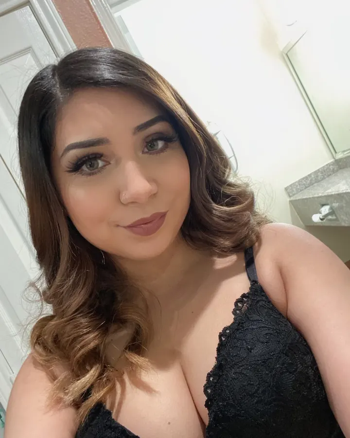 Femme prenant un selfie devant un miroir, portant un haut noir à boutons et une jupe à pois blancs, dans une salle de bain.