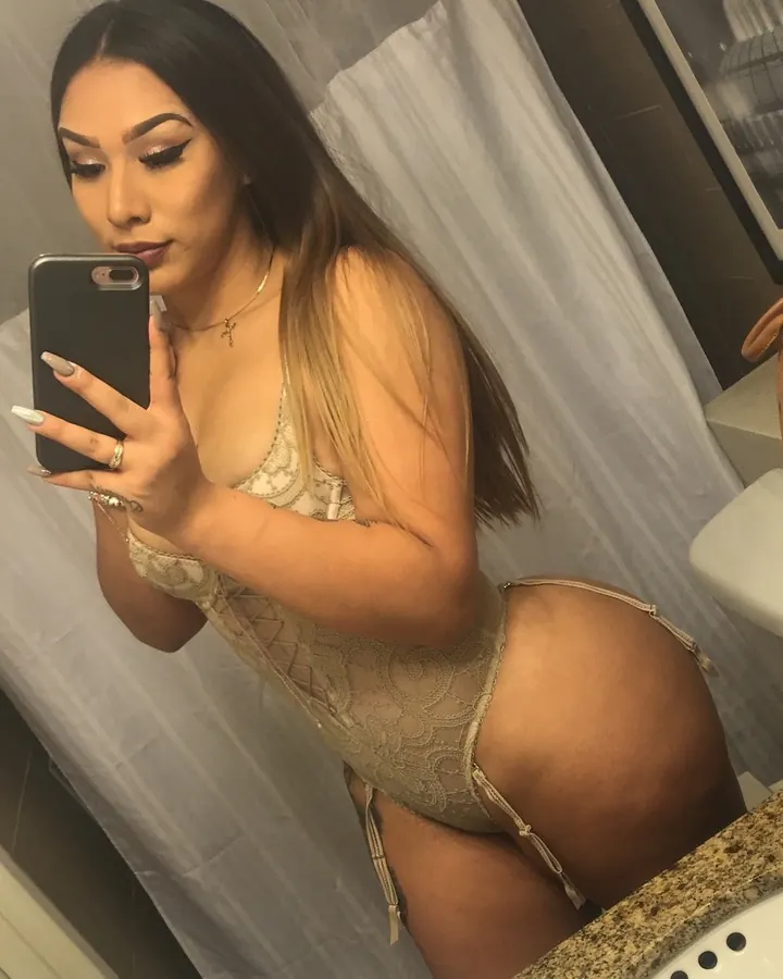 Femme aux cheveux longs, vêtue d'un body transparent beige, prenant un selfie dans un miroir, avec des bijoux aux doigts.