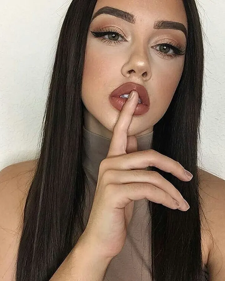 Femme aux cheveux foncés, vêtue d'un haut gris et d'un short en jean, allongée sur un lit blanc, avec un regard doux.