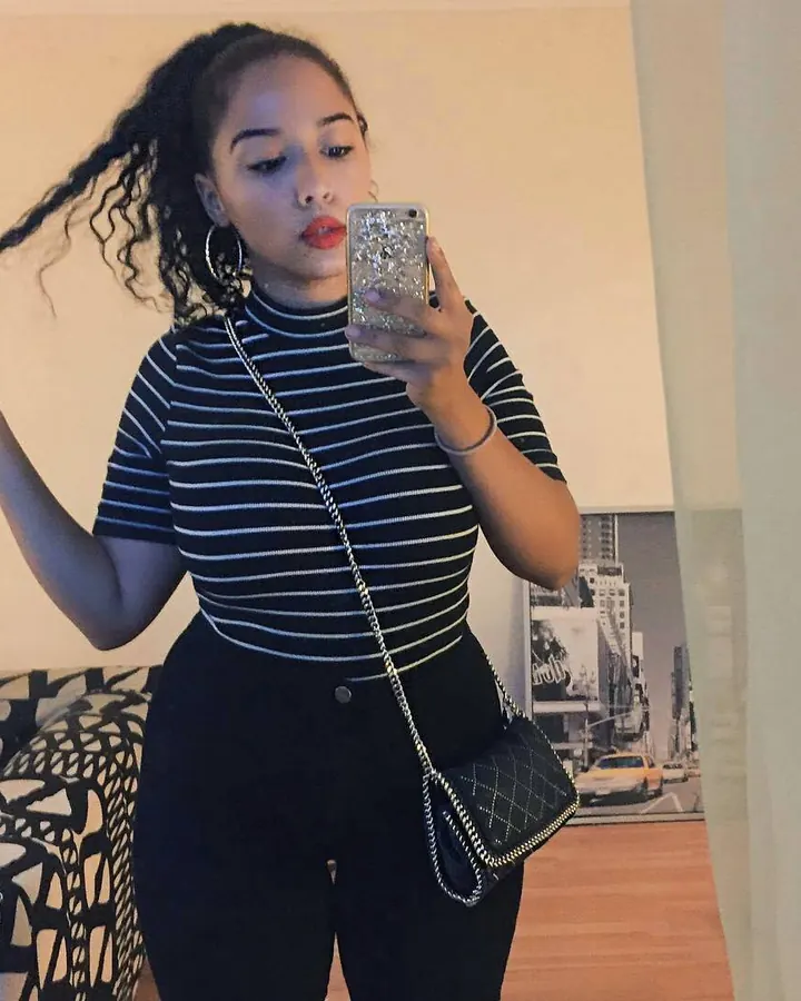 Femme debout, portant un haut noir et un jean bleu, les mains sur ses seins, tenant un téléphone doré pour se prendre en photo, avec un collier et une montre blanche.