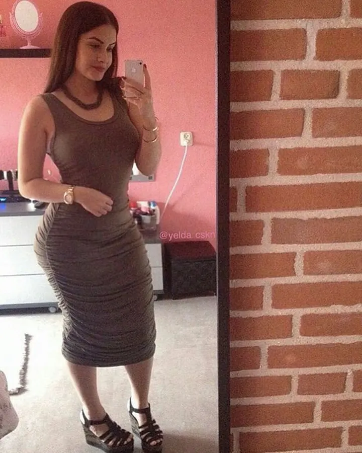 Femme en robe grise ajustée, prenant un selfie dans un miroir, dans une chambre avec mur rose.