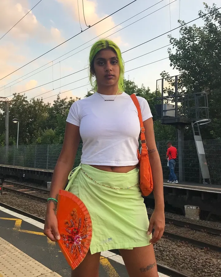 Femme en tenue colorée, t-shirt blanc, jupe vert citron, sac orange, tenant un éventail, sur un quai de train.